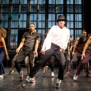 Group of professional dancers performing a high-energy routine on stage, led by a dancer in a white shirt and black hat.