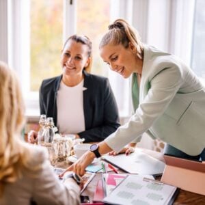 Three professional women collaborating during an event planning meeting, smiling and reviewing notes at a table.